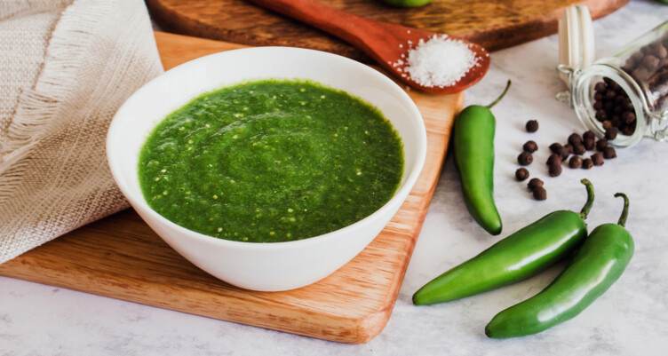 homemadegreen hot sauce in white bowl next to peppers and seasonings