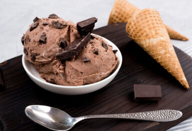 Scoops of chocolate ice cream in a bowl with a spoon and two waffle cones surrounding the dish.