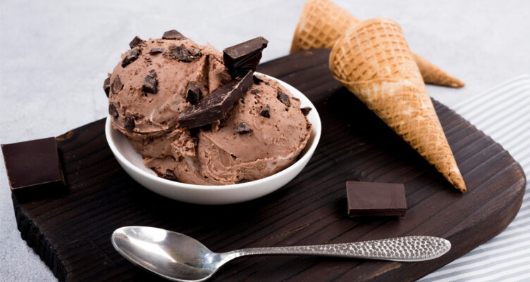 Scoops of chocolate ice cream in a bowl with a spoon and two waffle cones surrounding the dish.
