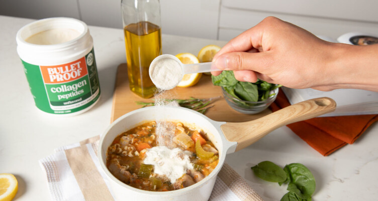 Hand pouring a scoop of Bulletproof Unflavored Collagen Peptides into a pot full of soup ingredients in a kitchen setting.