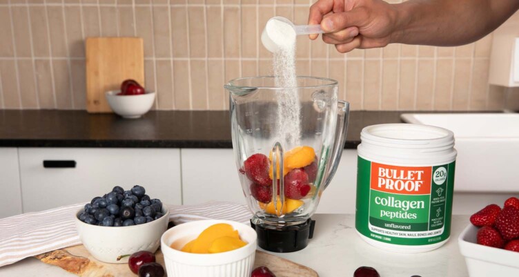 Hand pouring a scoop of Bulletproof Collagen Peptides into a blender filled with various fruits in a kitchen setting.