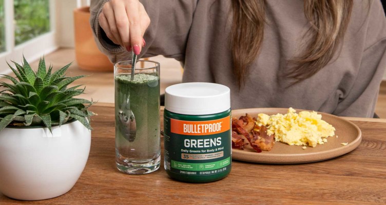 A hand stirring Bulletproof Greens into a glass of water alongside a plate of bacon and eggs