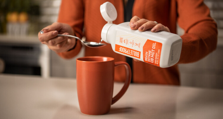 Woman in orange jacket pouring Brain Octane Oil onto a spoon above orange mug