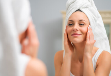 Woman looking in mirror and touching face while smiling and wearing a white towel on her head.