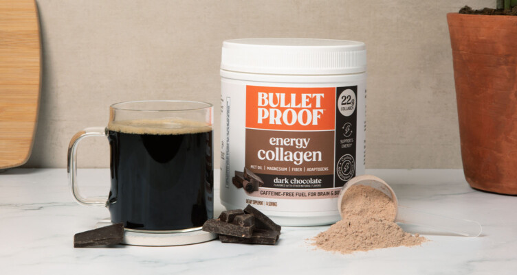 Tub of Bulletproof Dark Chocolate Energy Collagen sitting next to coffee mug with chocolate chunks and scoop scattered around.