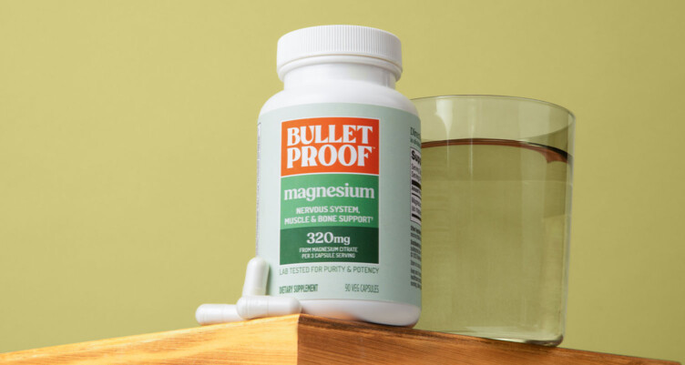 Bottle of Bulletproof Magnesium supplement sitting on a wooden pedestal with glass of water and pills next to it on a green background.