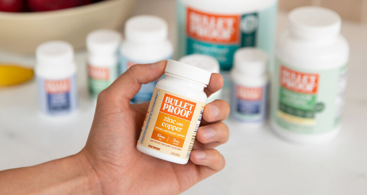 Hand holding a bottle of Bulletproof Zinc with Copper supplement with other bottles of supplements blurred out in the background.