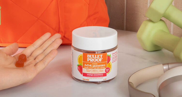 Hand holding a serving size of Bulletproof ADK Gummy Vitamin supplements with the tub sitting next to a gym bag, headphones and dumbbells.