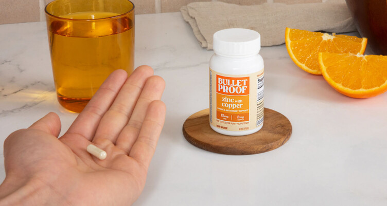 Hand holding a bottle of Bulletproof Zinc with Copper supplement with other bottles of supplements with a glass of water in the background.
