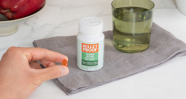 Hand holding a bottle of Bulletproof Vitamin A-D-K supplement with other a glass of water in the background.