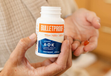 Woman holding Bulletproof A-D-K Vitamins bottle in one hand & the orange capsule in the other hand close to the chest
