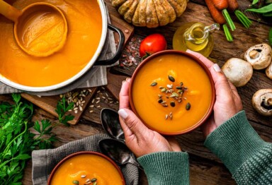 An overhead shot of a woman's hands holding a bowl of pumpkin soup with fall themed ingredients on a tablescape.