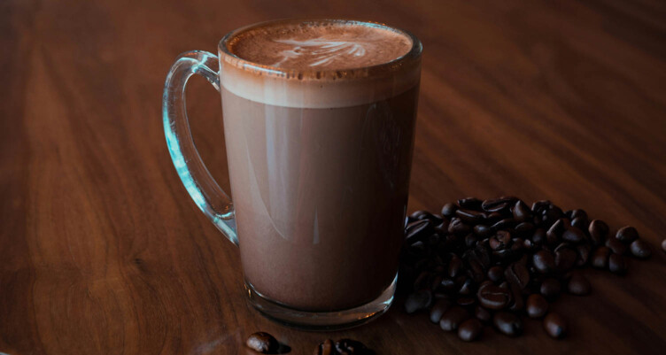 Clear mug filled with a frothy hot chocolate sitting on wooden surface and surrounded by coffee beans