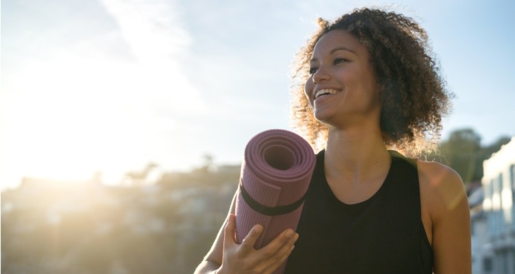 Fit woman smiling while holding a yoga mat at the beach.