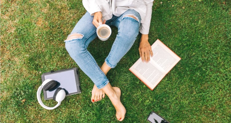 Woman sitting on the grass drinking coffee and reading a book.