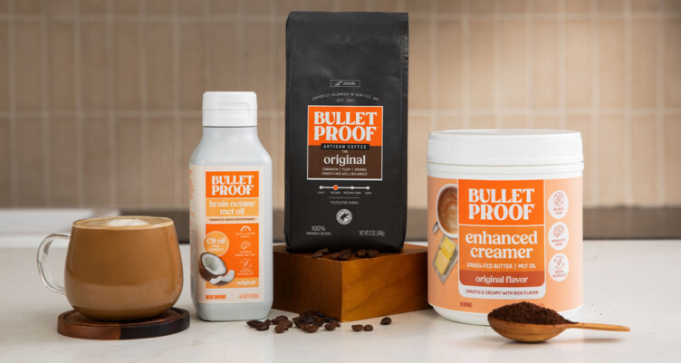 Bulletproof Original Ground Coffee bag, bottle of Brain Octane C8 MCT oil, tub of Original Enhanced Creamer and mug of coffee on kitchen counter with scoop of coffee grounds.