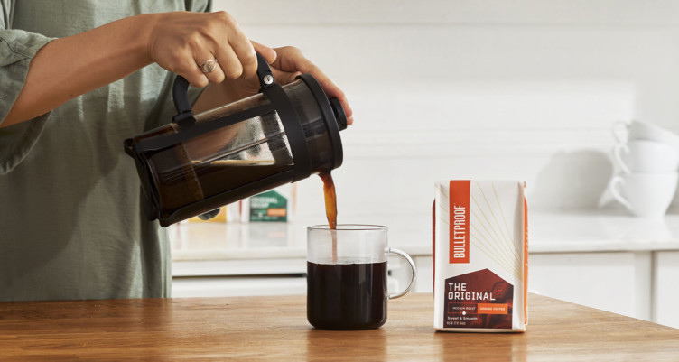 A hand pouring a French press of coffee into a mug next to a bag of Bulletproof coffee beans