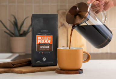 Hand pouring Bulletproof coffee from a French press with a bag of Bulletproof Original coffee sitting next to it on a cutting board in a kitchen setting.