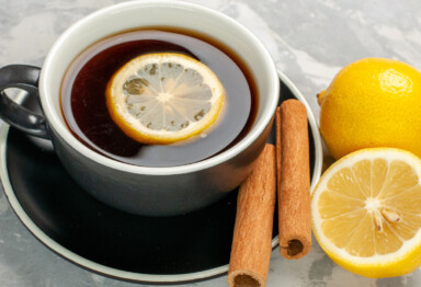 Mug fill with coffee with a lemon slice floating on top and cinnamon sticks next to mug on countertop.