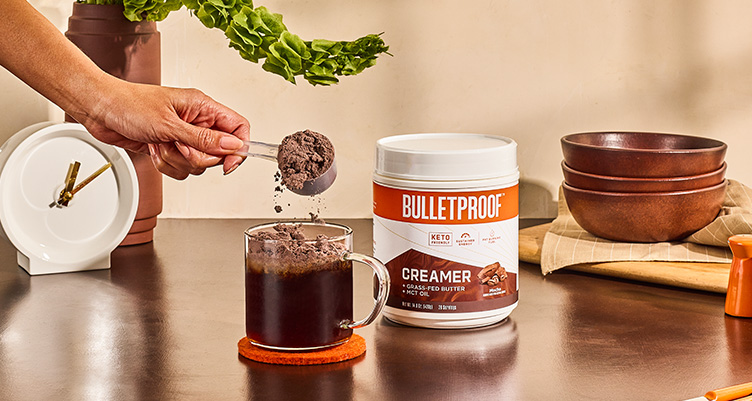 a hand of coffee pours a scoop of mocha bulletproof creamer into a cup of coffee on a table with various kitchen instruments surrounding