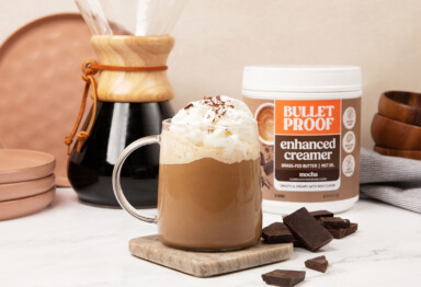 Mug filled with creamy coffee topped with whip cream and chocolate shavings, sitting in front of Bulletproof Enhanced Mocha Creamer and chemex.