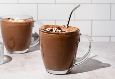 Two clear mugs filled with keto frozen hot chocolate drinks, toped with whip cream and chocolate chunks sitting against white tiled backdrop.