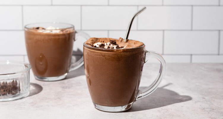 Two clear mugs filled with keto frozen hot chocolate drinks, toped with whip cream and chocolate chunks sitting against white tiled backdrop.