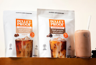Two bags of Bulletproof High Protein Iced Coffee in Original and Mocha flavor sitting on a pedastal next to a glass of iced coffee with a cream colored background.