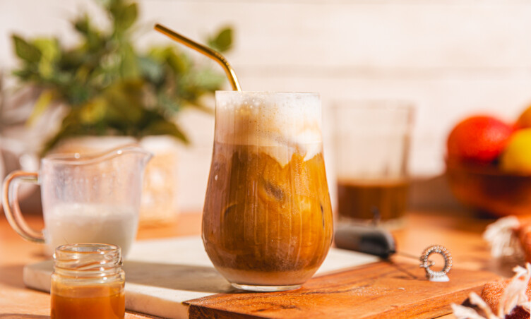 Glass of caramel iced coffee centered in frame, sitting on a cutting board surrounded by recipe ingredients and a frother.